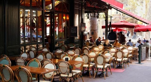 Paris café terrace with classic bistro chairs and red awnings, people dining outdoors on a sunny day.