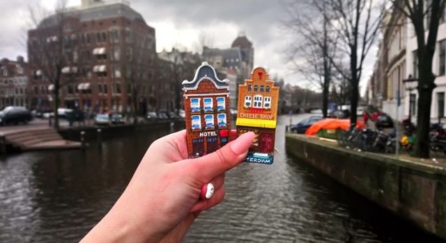 Hand holding Amsterdam souvenirs in front of a canal — iconic Dutch houses and waterway in the background.