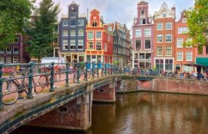 Amsterdam Food Guide: Best Local Eats, Markets & Dutch Classics “Amsterdam canal with colorful Dutch houses, bicycles on a bridge, and people walking in the background