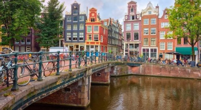 “Amsterdam canal with colorful Dutch houses, bicycles on a bridge, and people walking in the background