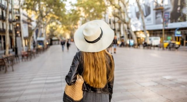 Woman in a wide-brim hat and light jacket walking along La Rambla, Barcelona in spring.