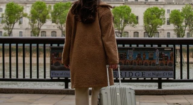 london-spring-what-to-wear-traveler A woman with a suitcase overlooking the Thames in early spring, wearing a warm coat in London.