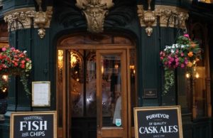 Best Pubs in London for a First Trip (Cozy, Historic, Riverside) Entrance of a traditional London pub with hanging flower baskets and signs for fish and chips and cask ales