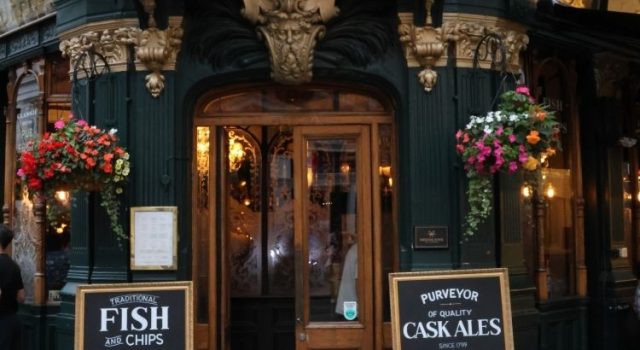 Entrance of a traditional London pub with hanging flower baskets and signs for fish and chips and cask ales