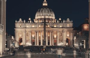 Rome at Night: Best Illuminations & Evening Walks (Map + Safety Tips) St. Peter’s Basilica illuminated at night with the obelisk in St. Peter’s Square and small crowds in motion blur