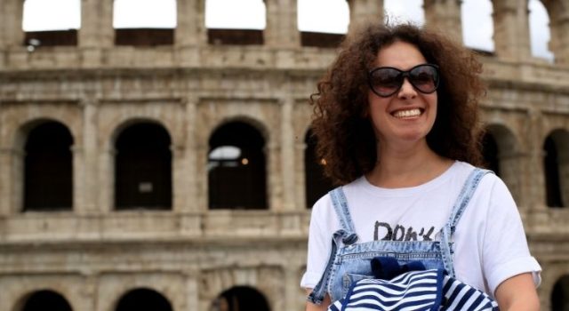 Woman in a T-shirt and denim overalls with sunglasses in front of the Colosseum in Rome