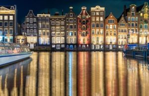 Where to Stay in Amsterdam: Best Neighborhoods for First-Time Visitors, Couples & Budget Travelers Night view of Amsterdam canal houses along Damrak, with colorful lights reflecting on the water.