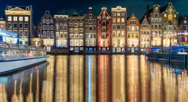Night view of Amsterdam canal houses along Damrak, with colorful lights reflecting on the water.
