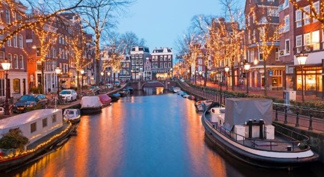 Amsterdam canal at dusk with illuminated trees, boats, and traditional Dutch houses—iconic cityscape for first-time visitors.