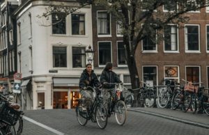 Amsterdam Packing & Weather Guide: What to Wear & Bring for Every Season Two women cycling on an Amsterdam street on a cloudy day, showing typical local clothing suitable for the city’s weather.