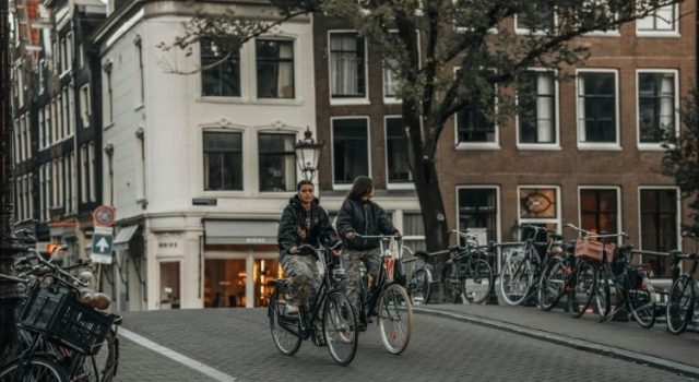 Two women cycling on an Amsterdam street on a cloudy day, showing typical local clothing suitable for the city’s weather.