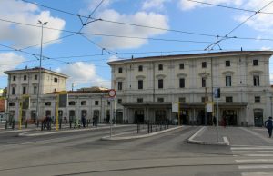 Rome Metro to Trastevere: The Station That Doesn’t Exist (And How to Get There) Front view of Roma Trastevere railway station in Rome, showing the historic cream-colored building, tram lines overhead, and people walking near the entrance.