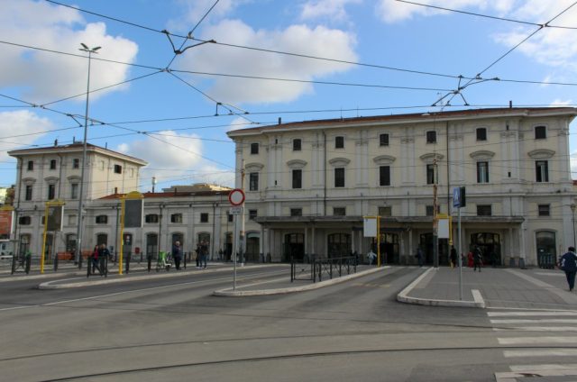 Front view of Roma Trastevere railway station in Rome, showing the historic cream-colored building, tram lines overhead, and people walking near the entrance.