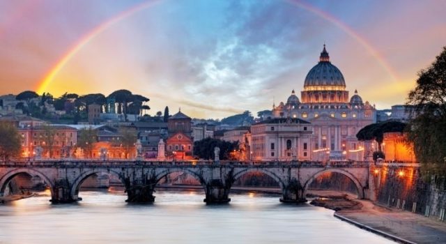 Scenic view of Rome at sunset with the Tiber River, an illuminated historic bridge, St. Peter’s Basilica in the background, and a rainbow arching across the sky.