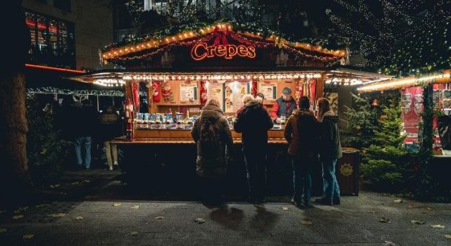 People ordering crêpes at a cozy outdoor market stall in Geneva during winter, with festive lights and a warm atmosphere.